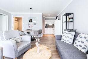 A modern living room with a grey sofa, a grey armchair, and a round coffee table. There is a dining area with a table and chairs, and a kitchen in the background. - Deluxe Baltic Stay für 6 Personen (Travemünde)