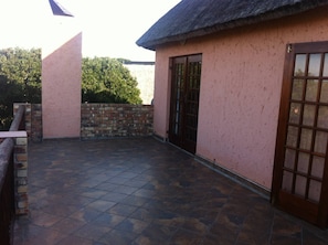 A covered patio area with a tiled floor, a brick wall, and a glass door leading to a building with a pink exterior. - Green Fountain Villa 5 (Port Alfred)
