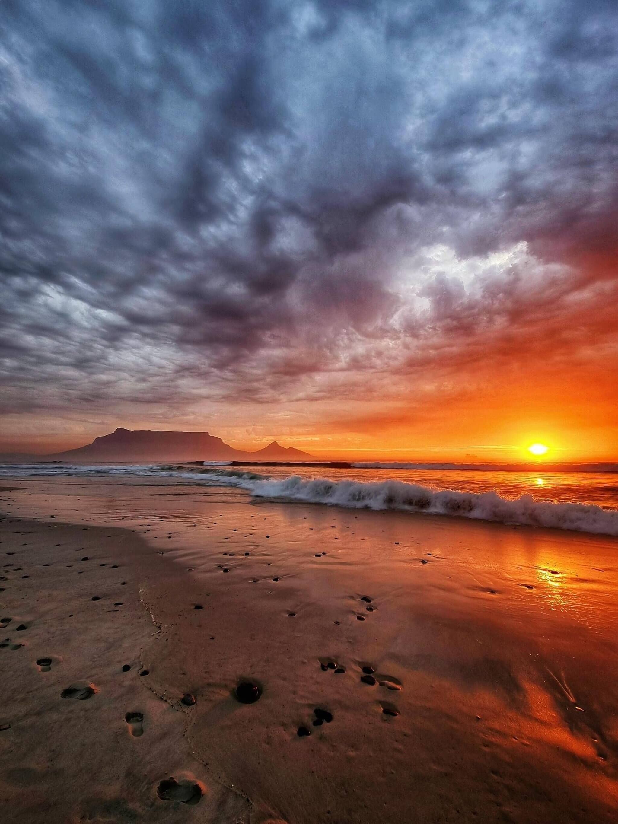 A beach with footprints leading towards the ocean, waves approaching the shore, and a sunset in the background.