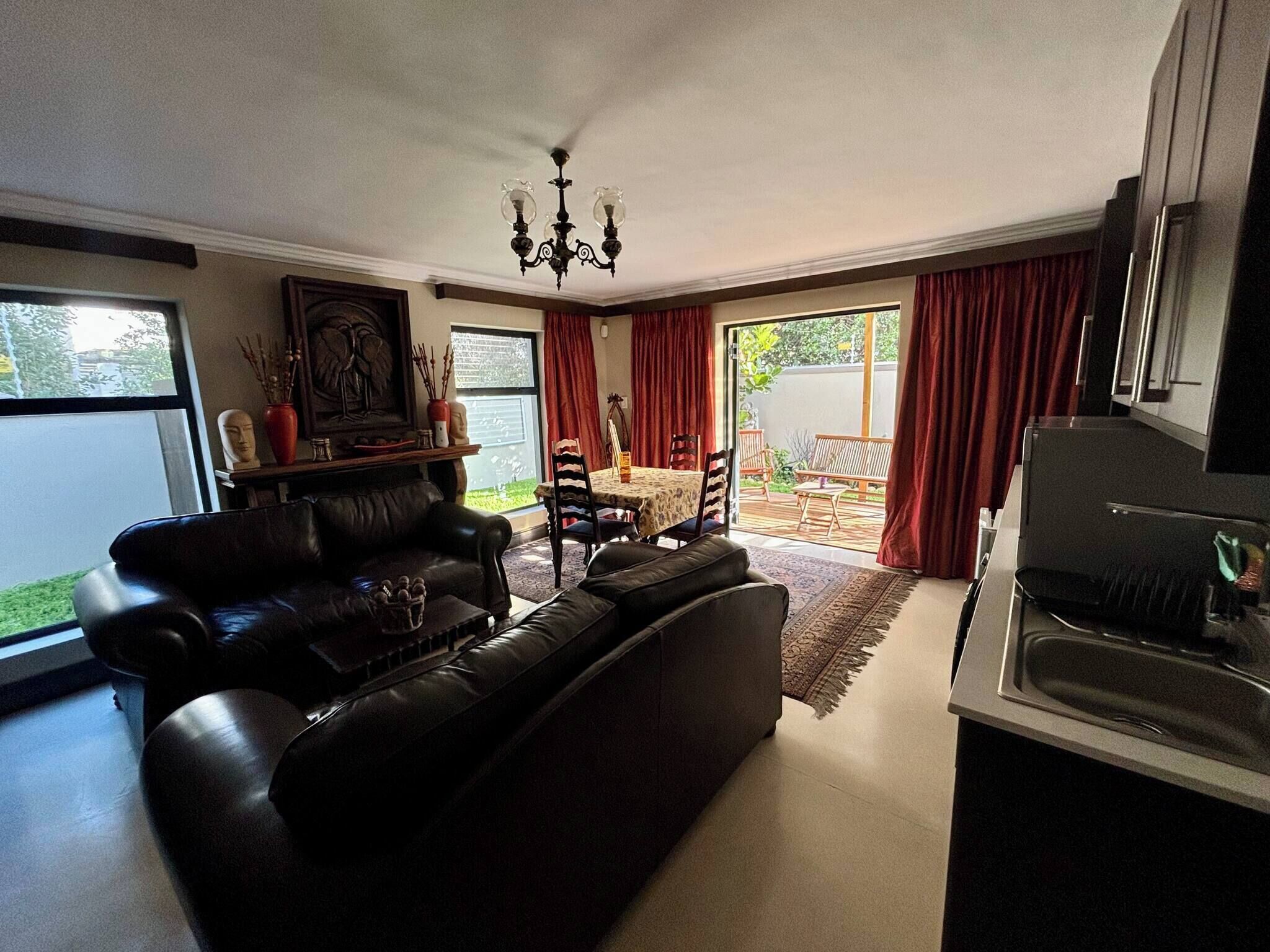 A living room with a black leather sofa, a dining table with chairs, a chandelier, and a kitchen area with a grill.