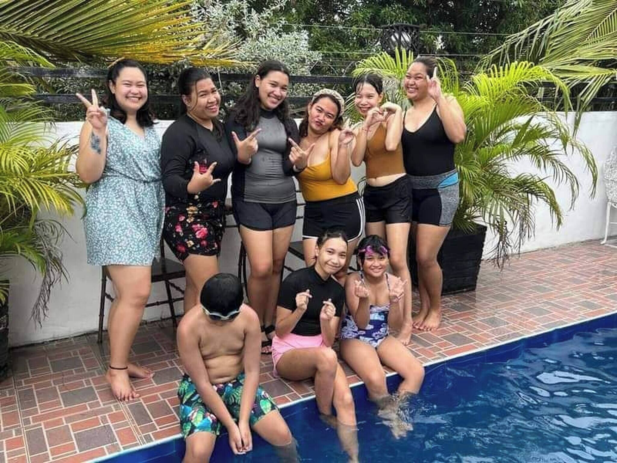 A group of people around a swimming pool with a tiled floor and palm trees in the background.