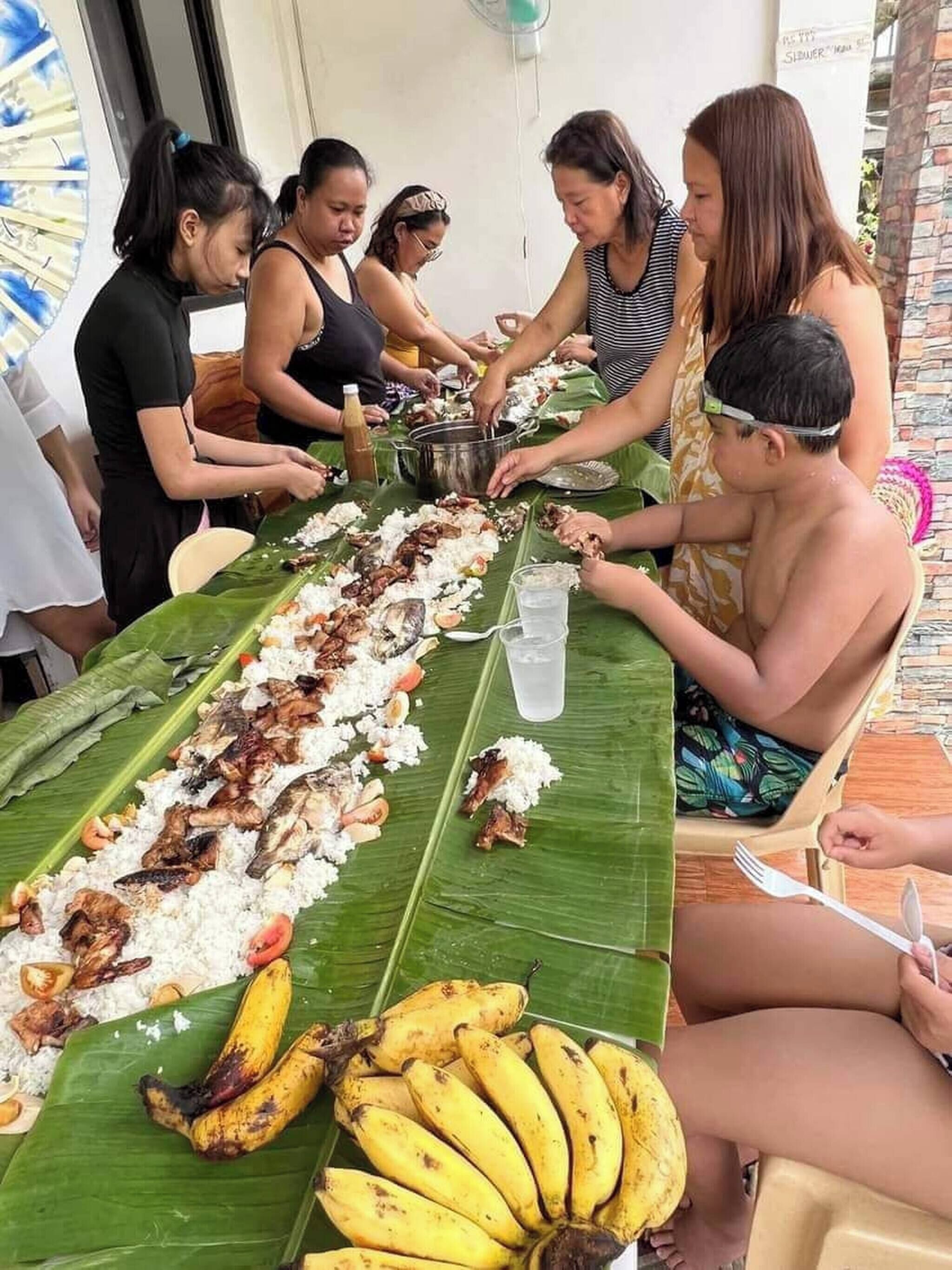 A group of people seated around a long table with food, including rice and bananas, in an outdoor setting.