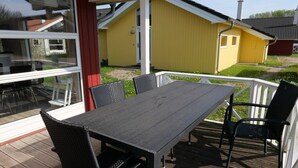 En terrass med ett bord och stolar, med utsikt över en gul byggnad och en grön gräsmatta.