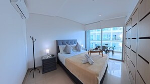 Habitación de hotel moderna con una cama grande, una mesita de noche con lámpara, una silla y vistas de la ciudad a través de amplios ventanales.