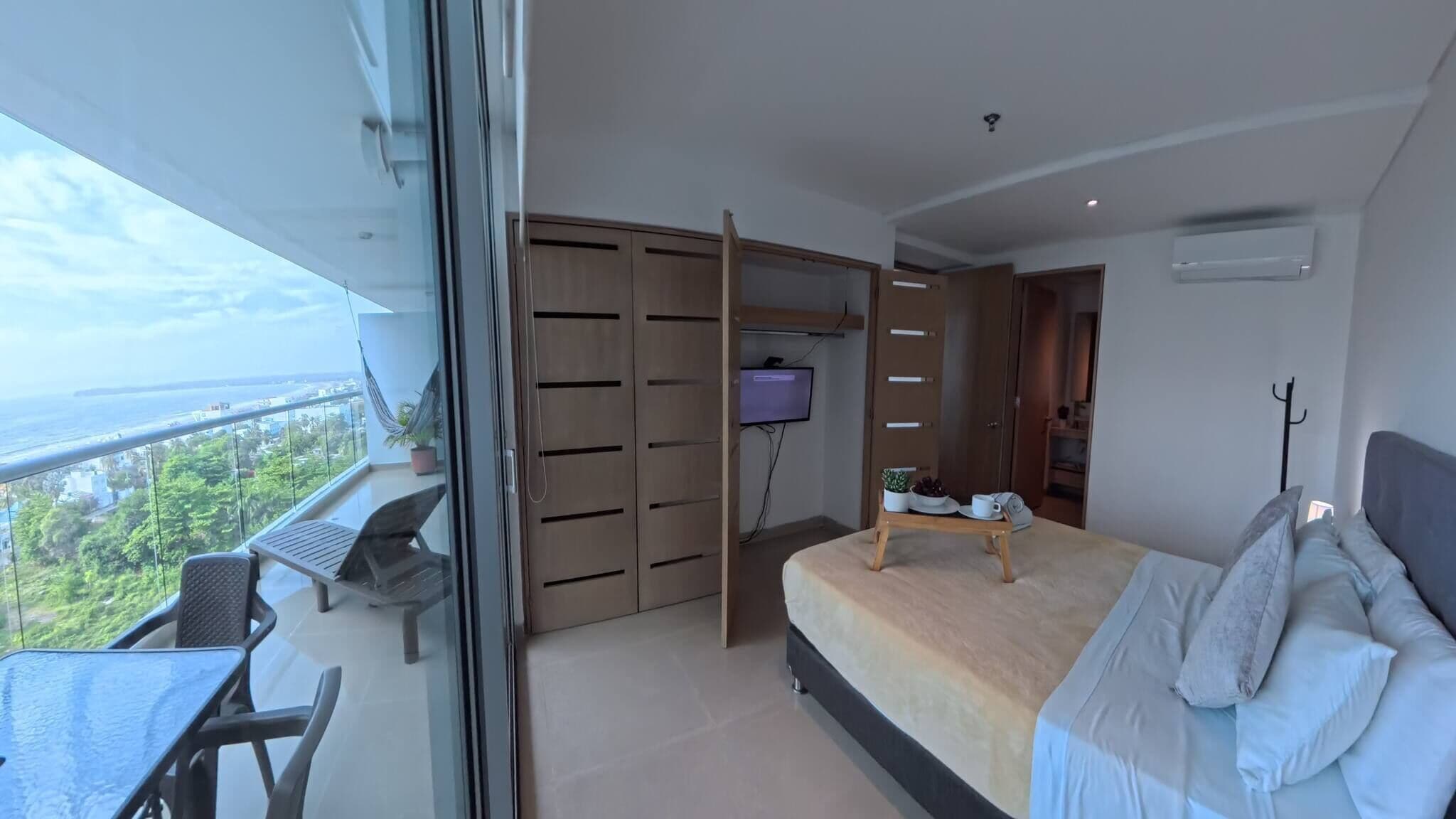 A bedroom with a bed, a desk, and a TV. There is a balcony with a view of the city and the sea.