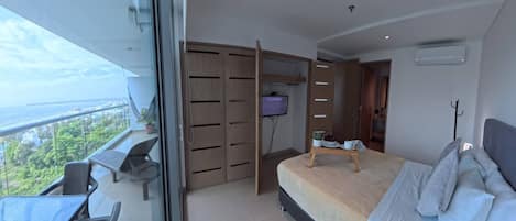 Un dormitorio con cama, escritorio y televisor. Tiene un balcón con vistas a la ciudad y al mar.