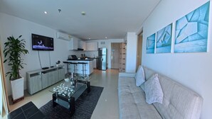 Una sala moderna con un sofá, una mesita de centro y un televisor. Se observa un área de cocina con un refrigerador y una barra.