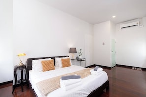 A neatly made bed with white linens and two decorative pillows, a bedside lamp, a small table with a lamp, and a wall-mounted air conditioner. - Spacious Heritage House Prime Location Family Stay (Khet Bang Rak)