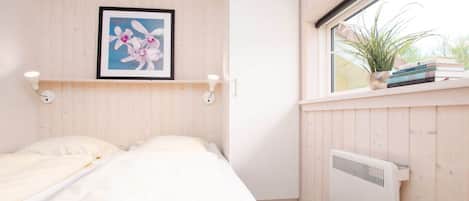 Un dormitorio con cama, un cuadro en la pared, un radiador y una ventana con planta y libros en el alféizar.