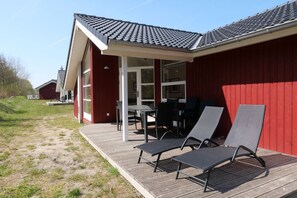 A red building with a covered patio area, a table, and two lounge chairs. - Grobenbrode-Haus mit Fußbodenheizung (Großenbrode)