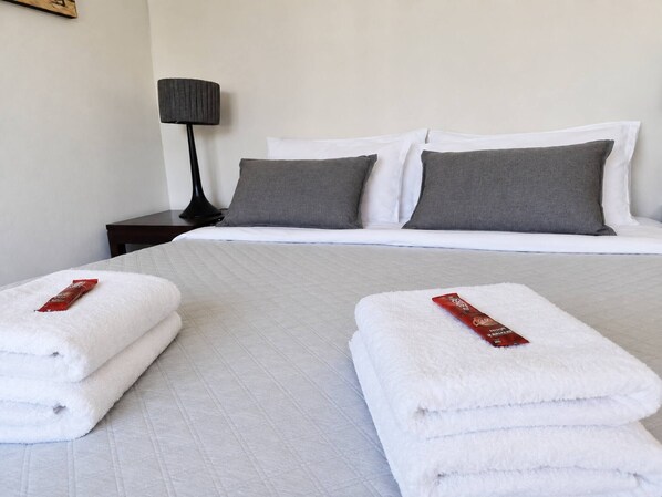 A hotel room with a neatly made bed, two pillows, a bedside lamp, and two folded towels with a red tag.