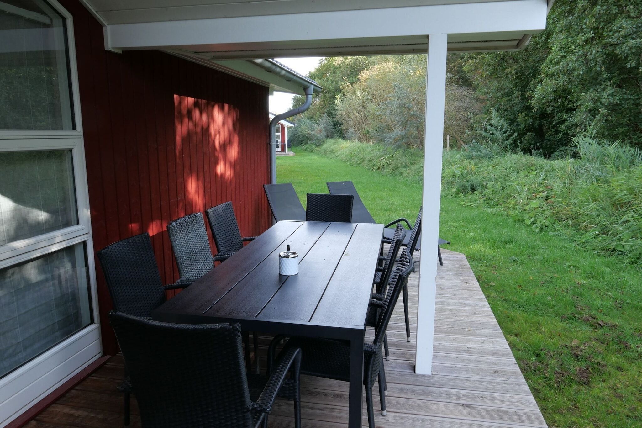 Un patio al aire libre con una mesa y sillas de madera, barandal blanco y vista a un jardín verde.