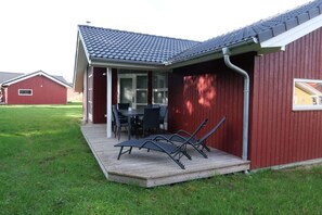 Een overdekt terras met een tafel en stoelen, een houten terras en een gebouw met een rode gevel en een pannendak.