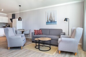 A modern living room with a grey sofa, two armchairs, a coffee table, and a painting of sailboats on the wall. - Apartment in Strand- und Parknähe (Travemünde)