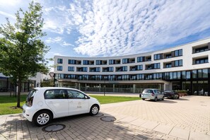 Un edificio moderno con techo plano, ventanas amplias y un acceso pavimentado con coches estacionados.