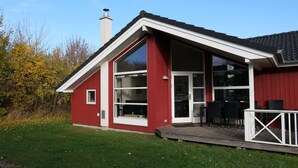 Ein rotes Gebäude mit großen Glasfenstern und einer Glastür, einem Schornstein und einer Holzterrasse mit Stühlen.