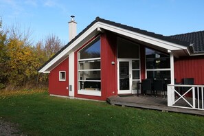Ein rotes Gebäude mit großen Glasfenstern und einer Glastür, einem Schornstein und einer Holzterrasse mit Stühlen.