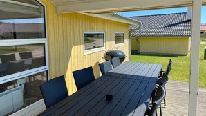 An outdoor patio area with a table and chairs, a grill, and a view of a road and buildings in the background. - Grobenbrode Escape für 10 (Großenbrode)