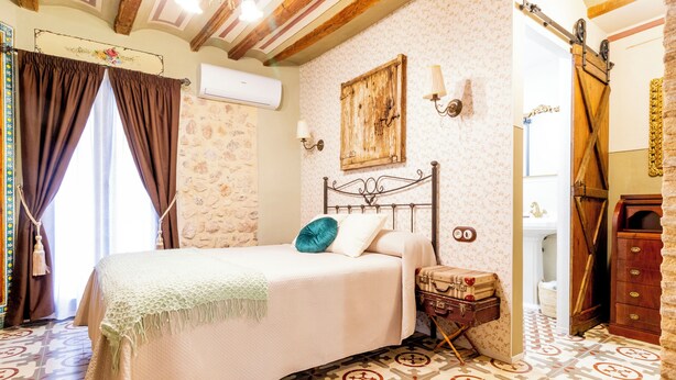 Un dormitorio con cama, mesita de noche, cómoda, lámpara de pared y ventana con cortinas.