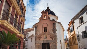 Una calle estrecha con una iglesia que presenta un campanario y una cruz, flanqueada por edificios con balcones y una palmera.