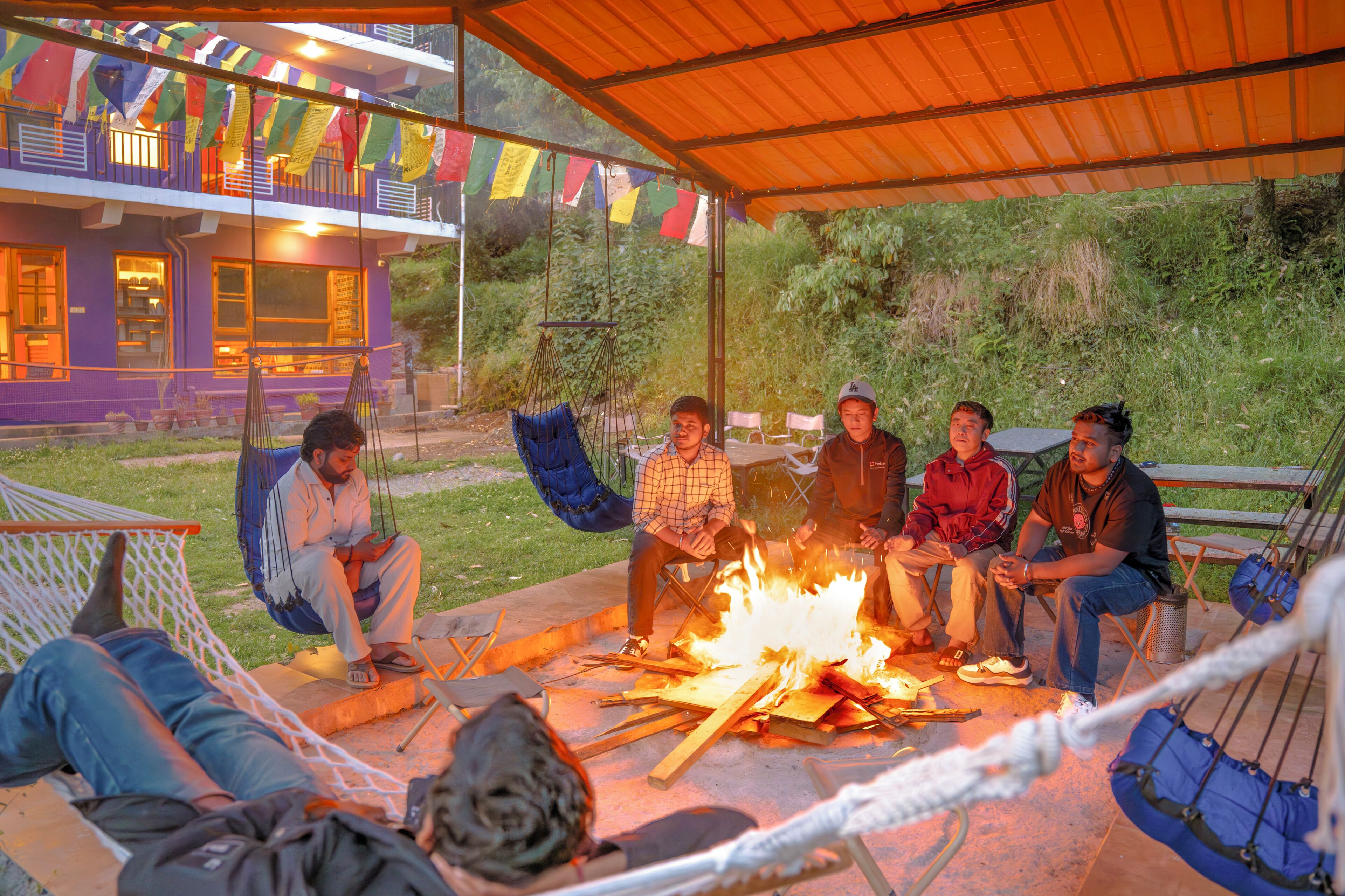 A group of people sitting around a campfire under a covered area with colorful flags.