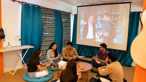 A group of people seated on bean bags in a room with a projector screen displaying a scene, surrounded by teal curtains and a whiteboard. - Loopin Mcleodganj  (Dharamshala)