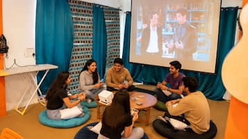 A group of people seated on bean bags in a room with a projector screen displaying a scene, surrounded by teal curtains and a whiteboard.