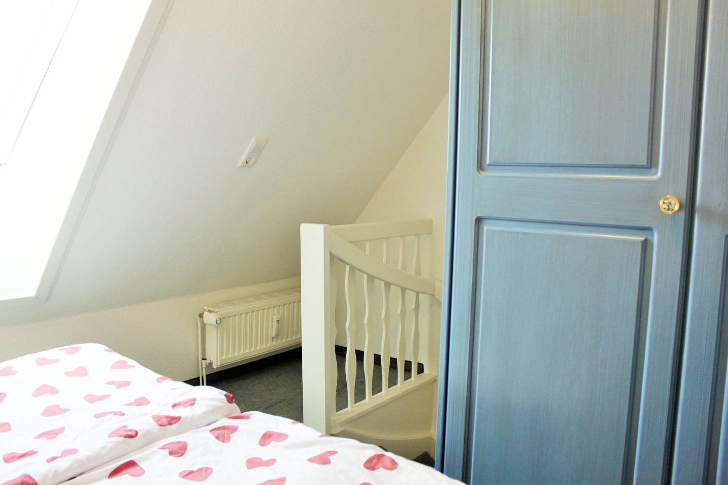 Een kamer met een schuine kap, een wit wiegje, een blauwe deur en een bed met een roggedeeltes beddegoed.