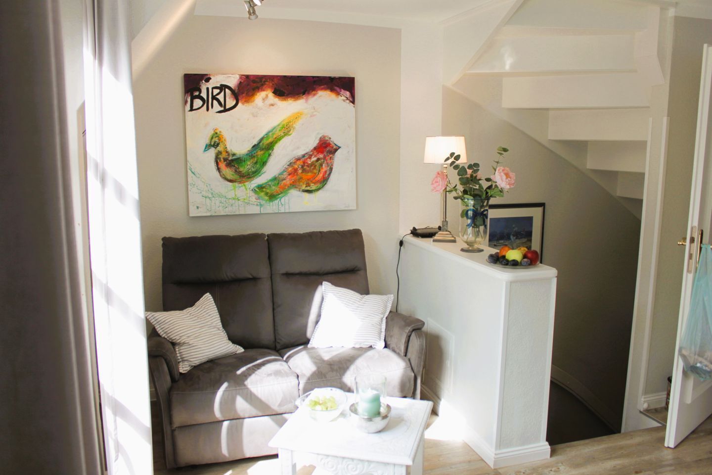 Een woonkamer met een bank, een schilderij, een lamp en een vaas met bloemen.