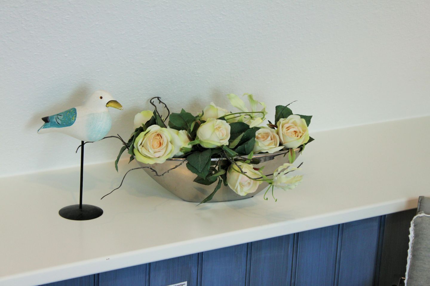 A decorative bird figurine on a stand next to a metal bowl containing white roses.