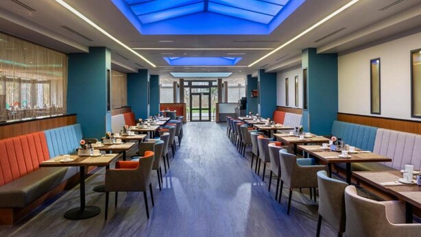 Comedor de hotel con cabinas y mesas, techo con tragaluces y vista a través de puertas de vidrio.