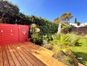 A wooden deck with a red gate, surrounded by a garden with various plants and a hedge. - The Good Life… (Fréjus)
