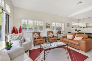 A spacious living room with a white sofa, two brown leather armchairs, a coffee table, and a dining area with a kitchen in the background.