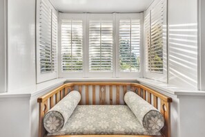 A bay window with white shutters, a wooden bench with patterned cushions, and a view of greenery outside.
