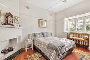 A bedroom with a bed, bedside table, lamp, and a window with blinds. - Bradman View | Bowral Garden and Fireplace Retreat (Bowral)