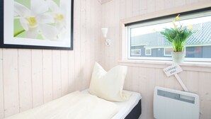 Habitació petita amb un llit individual, una finestra, un radiador, un quadre amb flors i un cartell que indica una zona d'estar.