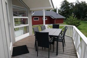 A covered outdoor area with a table and chairs, a red building in the background, and a white railing. - Küstenkomfort-Urlaub für 6 Personen (Großenbrode)