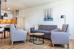 A modern living room with a grey sofa, two armchairs, and a coffee table. There is a painting of sailboats on the wall, a lamp, and a kitchen area in the background. - Meeresurlaub mit Wellness (Travemünde)