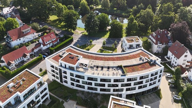 Um edifício moderno com disposição circular, cercado por vegetação e casas residenciais.