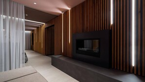 Um interior moderno com uma televisão de tela plana presa a uma parede escura, uma área de estar com um banco e um corredor com ripas verticais de madeira e um aparelho de luz.