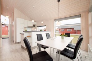 A modern kitchen with white cabinets and a microwave, a dining area with a white table and black chairs, and a view of a red building outside. - Charming Seaside Haven Oasis für 6 (Großenbrode)