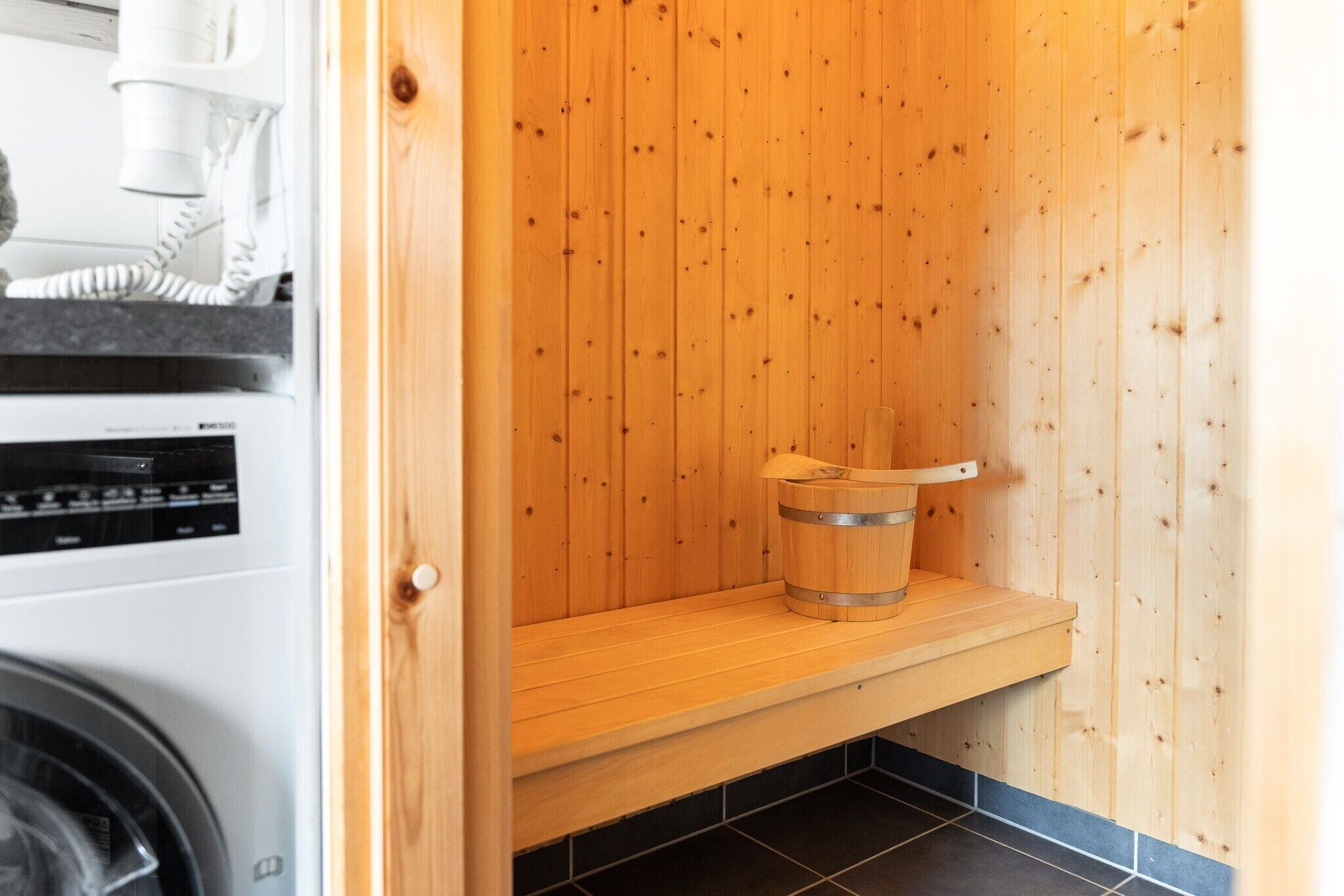 Un sauna avec un banc en bois et un seau, un lave-linge et un sol carrelé.