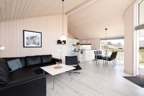 A modern interior with a black leather sofa, a white table, and a kitchen area with a dining table and chairs. - Küstenkomfort-Retreat für 6 Personen (Großenbrode)