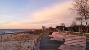 En træpromenade fører hen til en strand med bænke, et hegn og bygninger i baggrunden.