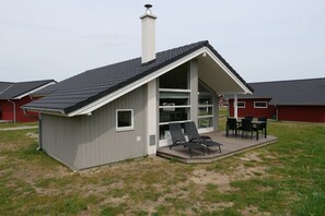 A small, single-story building with a sloped roof and a chimney. It has a large glass door and windows, a patio with outdoor furniture, and is surrounded by a grassy area. - Skandinavischer Rückzug für 6 (Großenbrode)