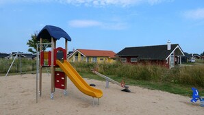 Un parque infantil con tobogán y columpios sobre arena, rodeado de casas.