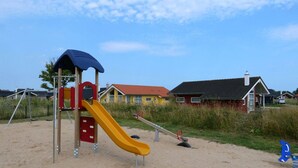 Un parque infantil con tobogán y columpios, rodeado de casas y una zona de arena.