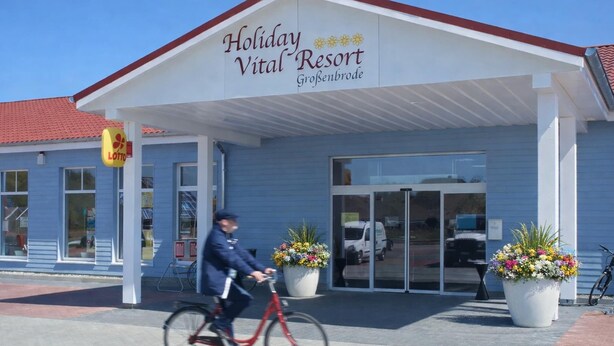 Edificio del Holiday Vital Resort con una entrada despejada, un letrero de Lotto y dos macetas grandes afuera.