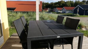 A wooden deck with a black table and chairs, surrounded by grass and a row of houses with red roofs. - Ruhiges Küstenretreat für 6 (Großenbrode)
