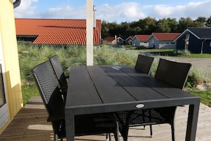 A wooden deck with a black table and chairs, surrounded by grass and a row of houses with red roofs. - Ruhiges Küstenretreat für 6 (Großenbrode)